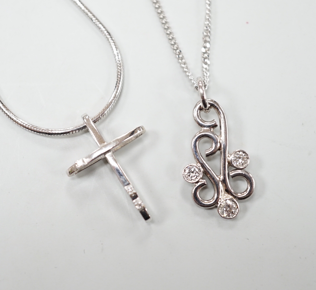 A modern 18ct white gold and three stone diamond set scroll pendant, 20mm, on an 18ct white gold chain, 39cm, gross weight 5.3 grams, together with a 585 white metal and diamond chip set cross pendant, on a 585 white met